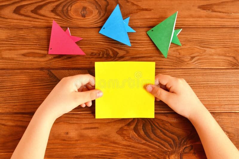 Child holds a paper square in his hands. Child making origami fish. Set of origami fish on a wooden table royalty free stock photography
