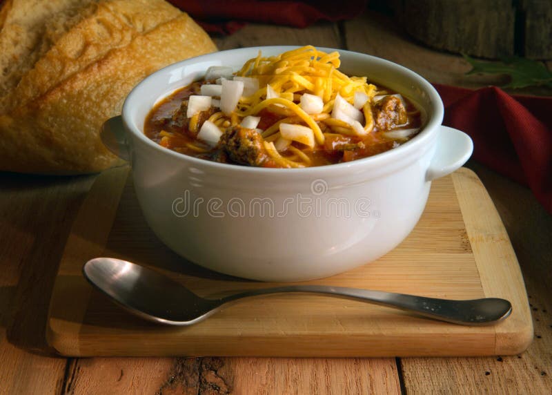 Chili in white crock with onions and cheddar cheese on rustic wood table. White crock of chili with raw onions and shredded cheddar cheese on a wood board on a royalty free stock image