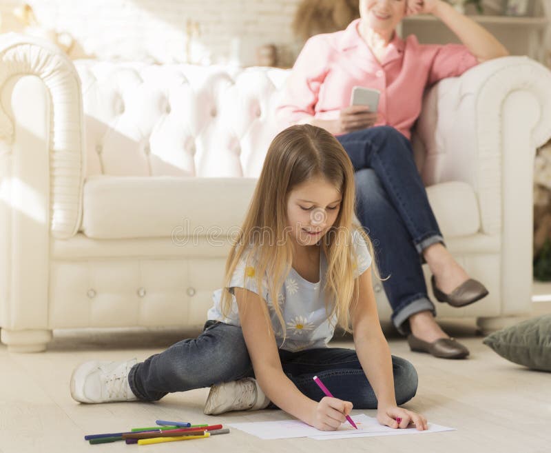 Cute girl drawing on floor while her granny using mobile phone at home. Lovely girl drawing on floor while her granny using mobile phone at home stock photography