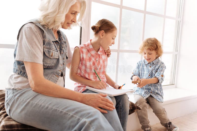 Cute pleasant girl looking at her brother. What have you brought. Cute pleasant nice girl sitting together with her grandmother and holding a drawing while stock photo