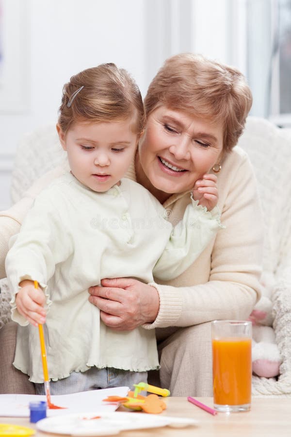 Cute small girl is painting with her grandparent. Pretty granddaughter is learning to paint with her grandmother. They are sitting on sofa and smiling. The women stock images