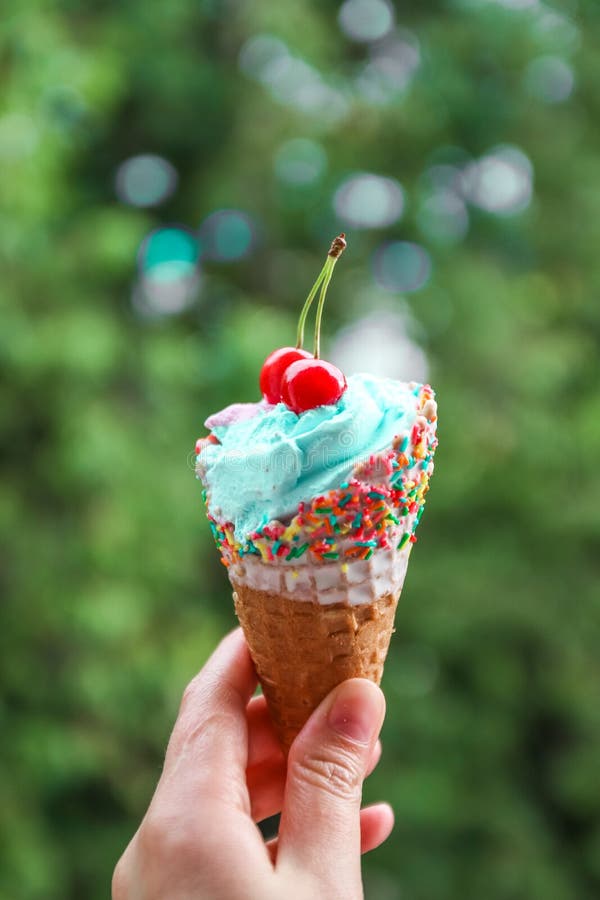 Female hand holds multi-colored ice cream with cherries in a waffle cone against blur background. Female hand holds multi-colored ice cream with cherries in a royalty free stock photo