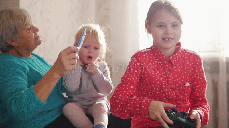 A grandmother combing her granddaughter`s hair. Another girl playing with joystick. Mid shot stock image