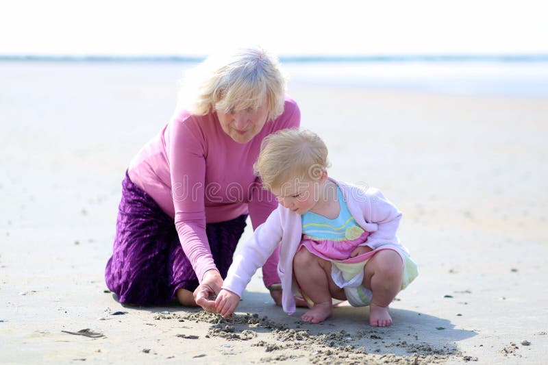 Grandmother and granddaughter playing together on the beach. Happy grandmother playing with her granddaughter, cute toddler girl, at the beach drawing on the stock photo