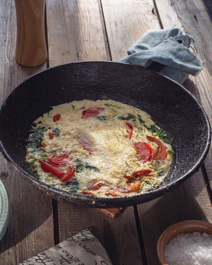 Omelet with tomatoes in a round cast iron pan on an old Board table, a napkin with a pattern and a large salt in a salt. Omelet with tomatoes in a round cast royalty free stock images