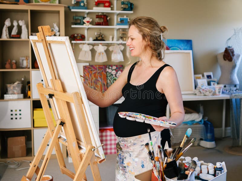 Portrait of smiling white Caucasian young pregnant woman drawing painting standing at easel in home studio. Art mental therapy, healthy happy lifestyle concept stock photo