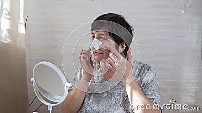 Woman in age, looking in the mirror, touches face with hands, takes care of skin at home, beauty concept. Anti-aging stock video footage